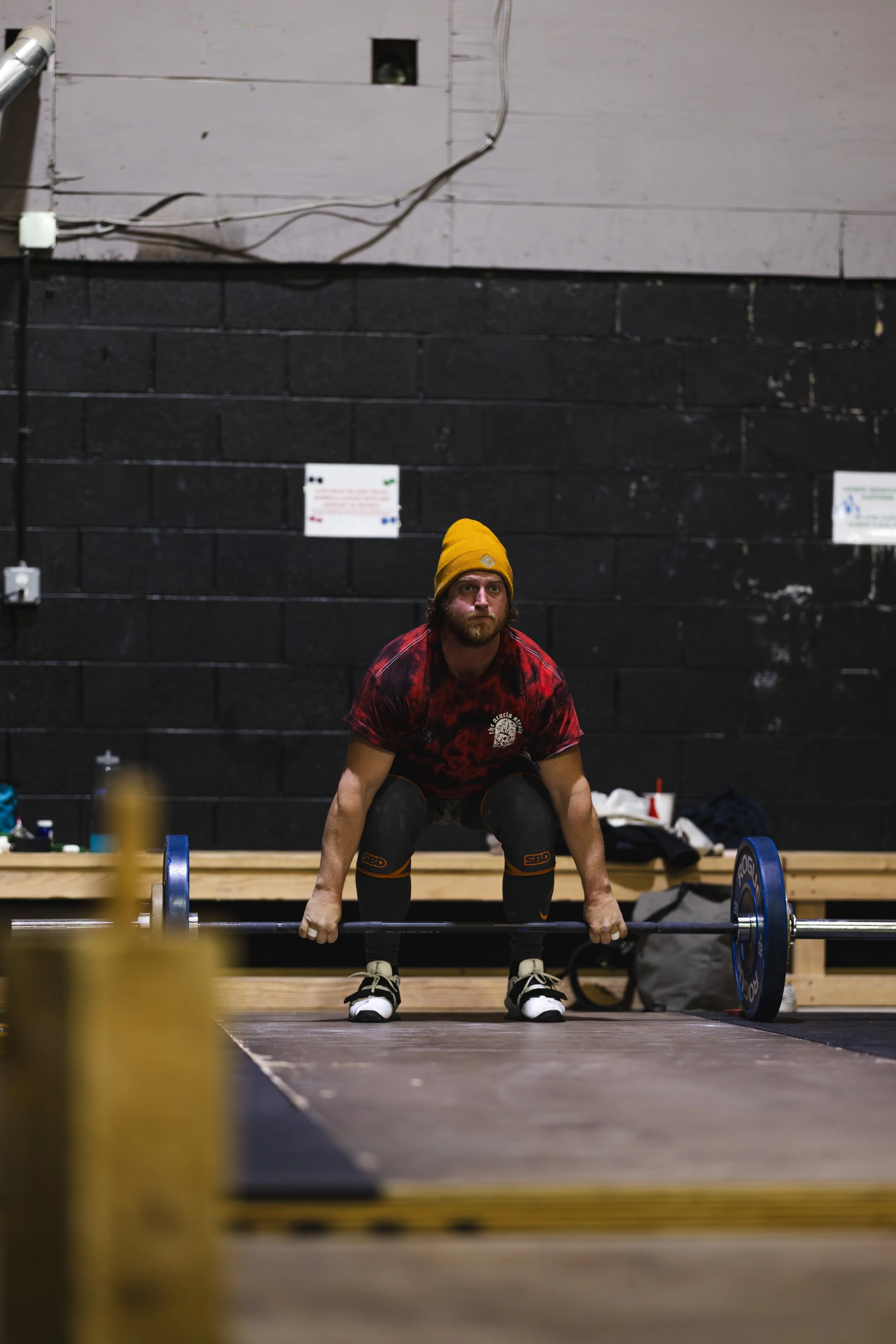 A man with a beard, wearing a yellow beanie, red tie-dye shirt, and black knee sleeves, is preparing to lift a barbell in an indoor gym with black walls.