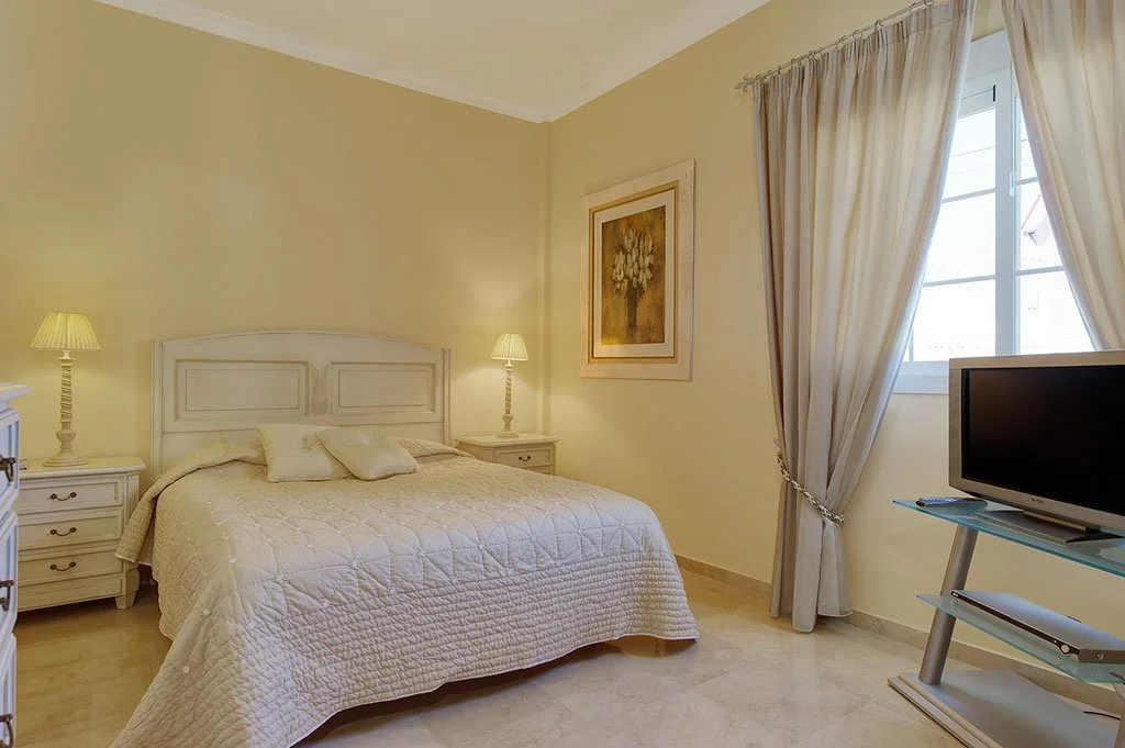 A cozy bedroom with light beige walls, a white wooden bed with a quilted cream bedspread, two matching nightstands with lamps, a framed floral painting on the wall, a window with cream curtains, and a flat-screen TV on a glass stand.