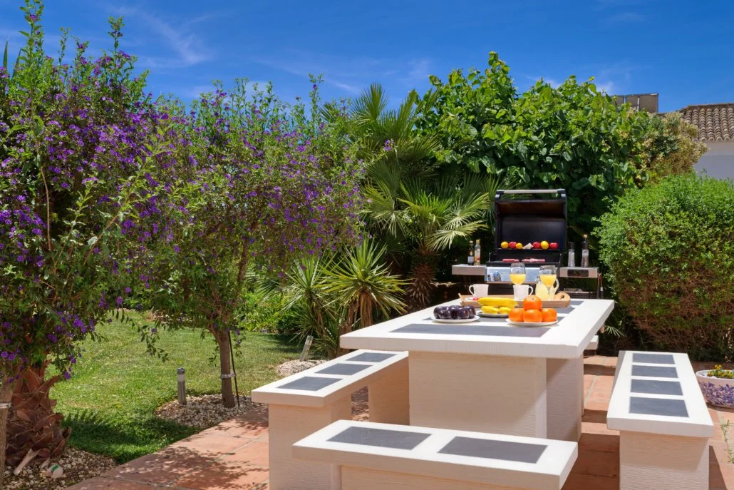 Outdoor patio with a white table and benches set for a barbecue, surrounded by green bushes and trees under a blue sky.