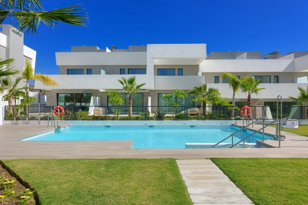 Residential apartment complex with a swimming pool, surrounded by palm trees and landscaping, under a clear blue sky.