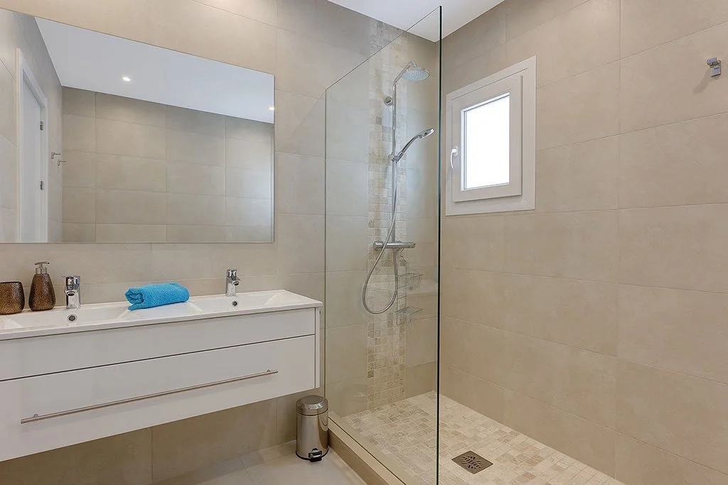Modern bathroom with a double sink vanity, large mirror, and walk-in shower with glass partition, beige tiled walls and floor, small window, and a waste bin on the floor.