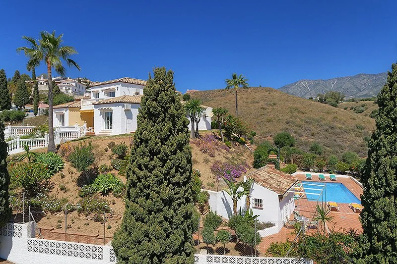 White houses on a hillside with a swimming pool and palm trees, under a clear blue sky with mountains in the background.