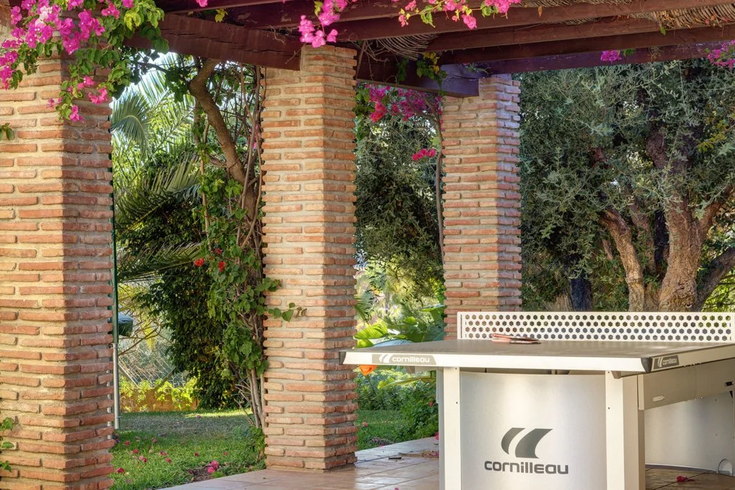 Outdoor patio with brick columns, surrounded by lush greenery and flowering vines, with a ping pong table on the right side.