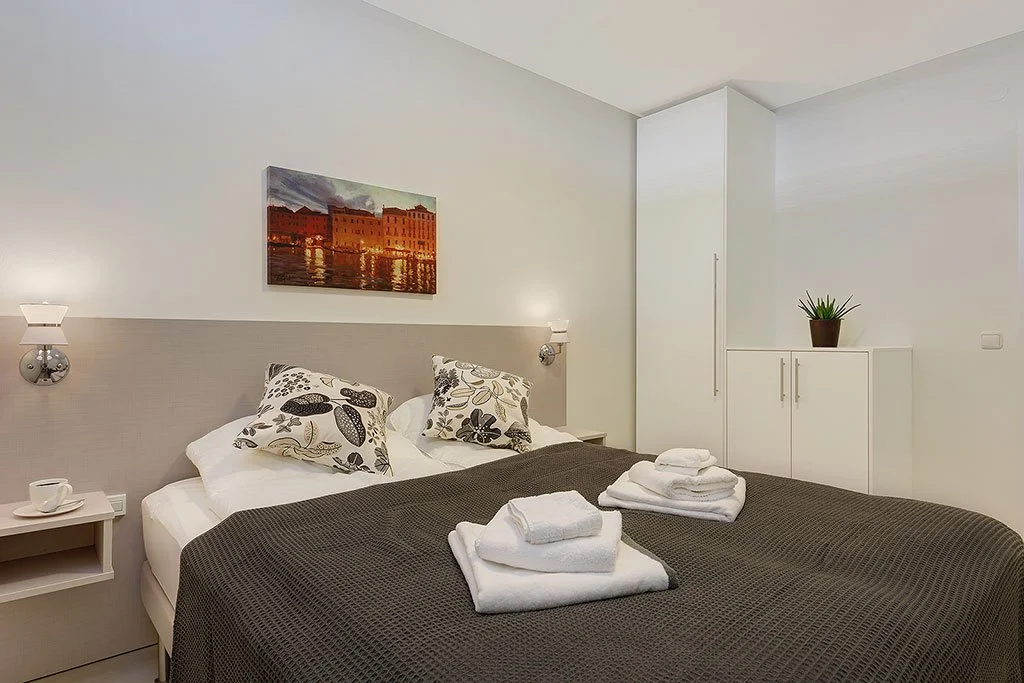 Hotel room with a king-sized bed, pillows with floral patterns, a dark blanket, folded towels on the bed, wall-mounted lights, a nightstand, a framed landscape painting above the bed, and a white cabinet with a potted plant.