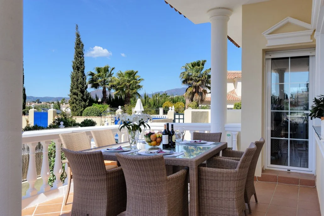 Outdoor dining area on a balcony with a glass table, six wicker chairs, and a view of palm trees and a blue sky.