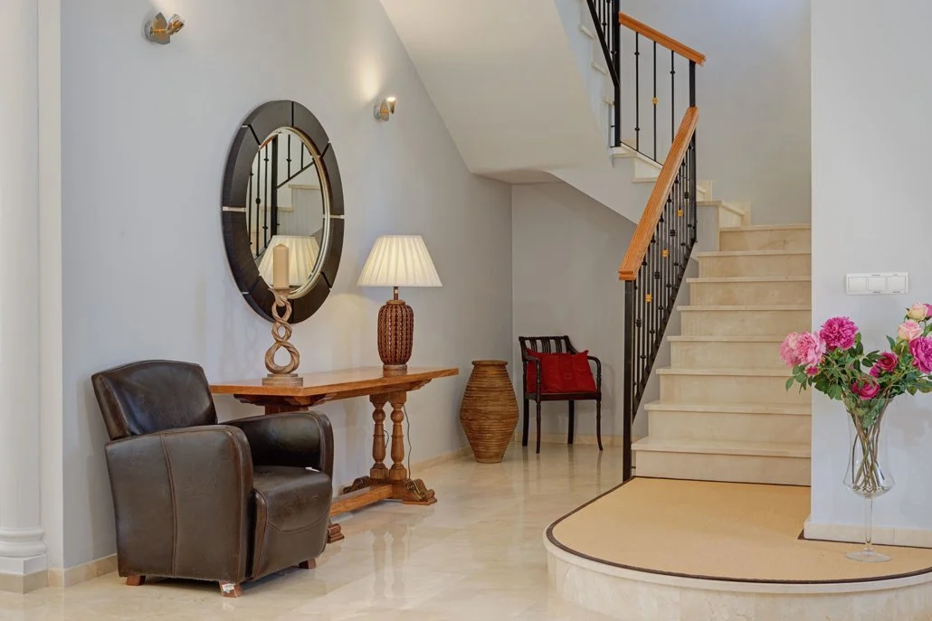 Interior of a home entryway with a staircase, a black leather armchair, a wooden side table with a lamp, a decorative mirror, a large vase, and a bouquet of pink flowers in a glass vase.