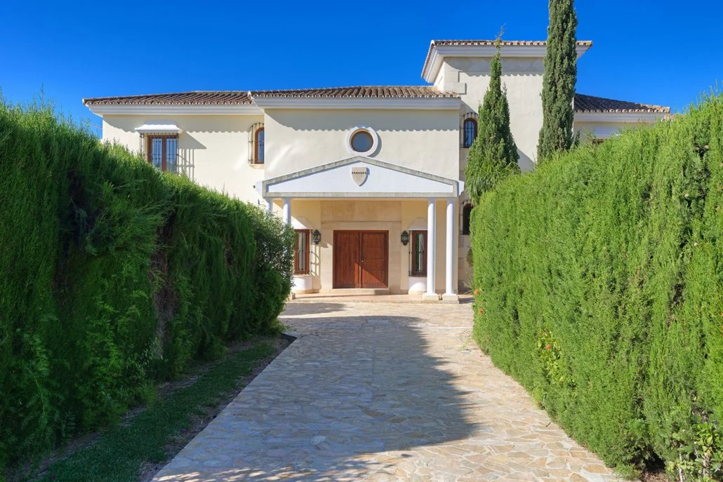 Front view of a large, white Mediterranean-style house with a cobblestone driveway, green hedges on both sides, and tall cypress trees in the garden, under a clear blue sky.