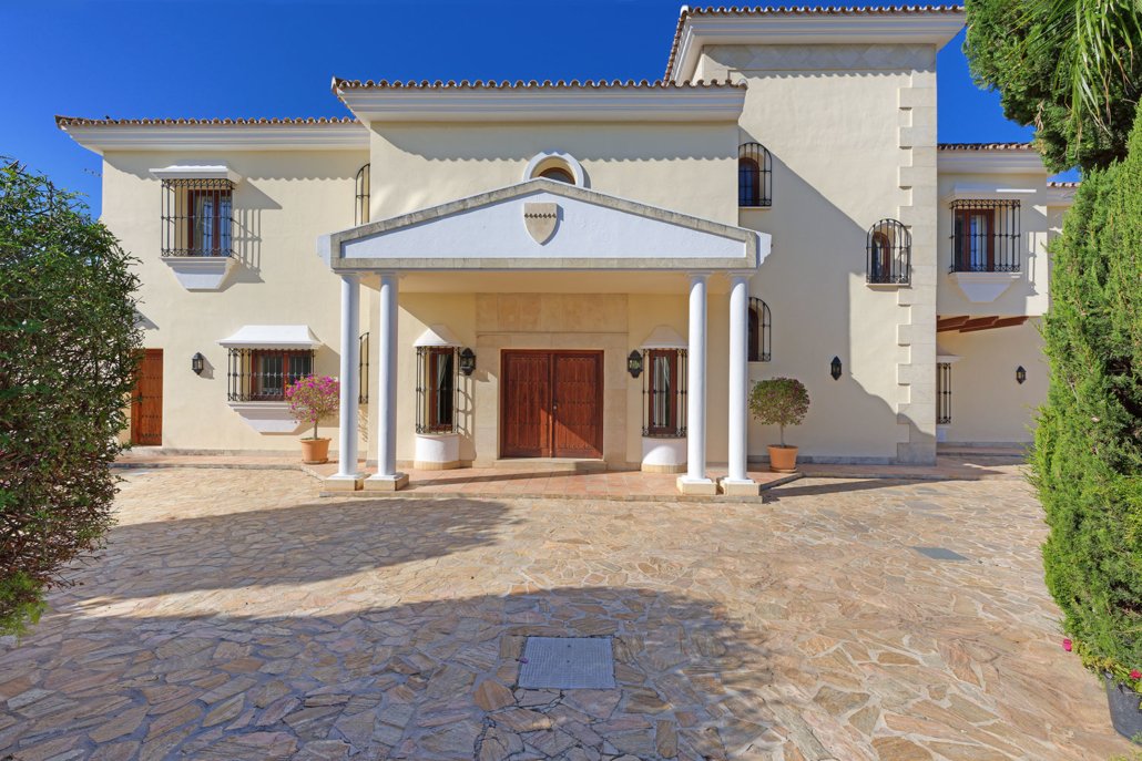 Front view of a large Mediterranean-style house with a stone driveway, white stucco walls, and a portico supported by columns.