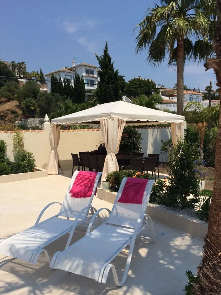 A backyard with two white sun loungers with pink towels, a white canopy with curtains, and a dining table with chairs underneath. There are palm trees and bushes, with houses on a hill in the background.