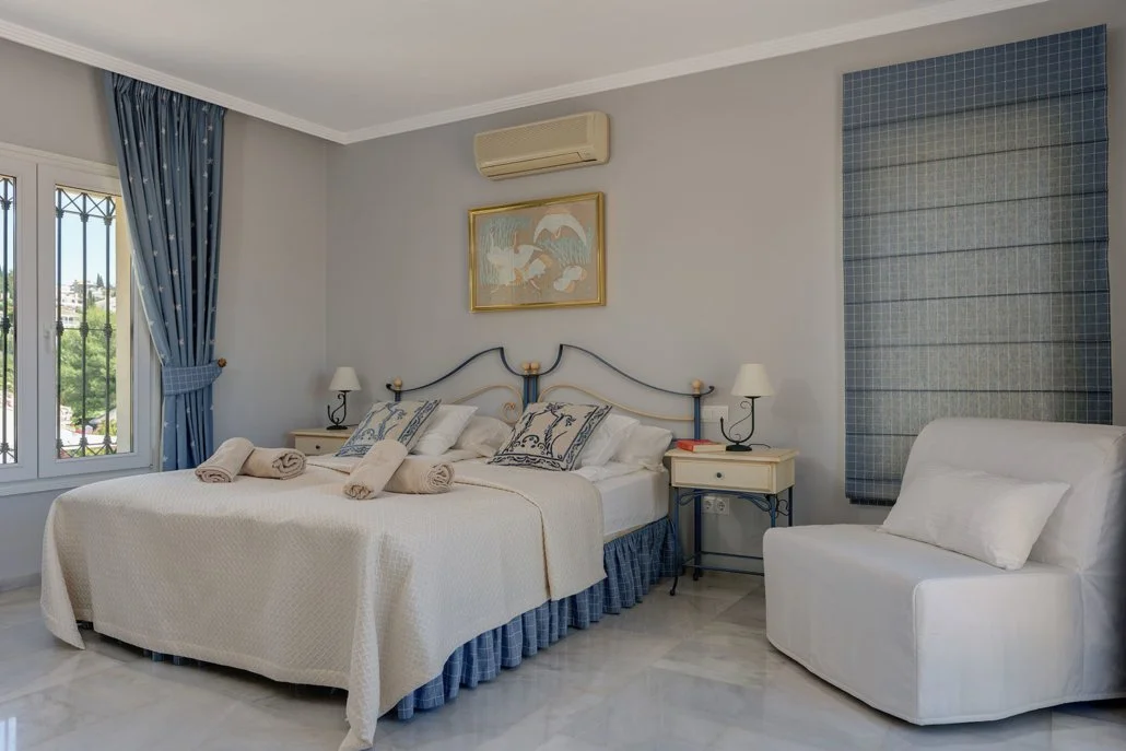 A cozy bedroom with a large bed, white bedding, and decorative pillows, flanked by two nightstands with lamps, blue curtains, a window with a view outside, a white armchair, framed artwork, and an air conditioning unit on the wall.