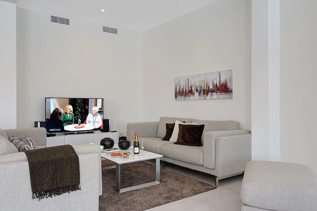 Modern living room with white walls, beige sofas, a television on a white stand, assorted black and brown pillows, a coffee table with a champagne bottle and snacks, and a cityscape painting on the wall.