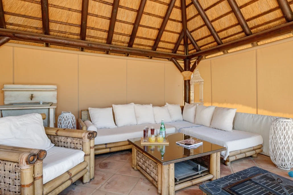 A tropical-themed lounge area with wicker furniture, white cushions, and a thatched roof.