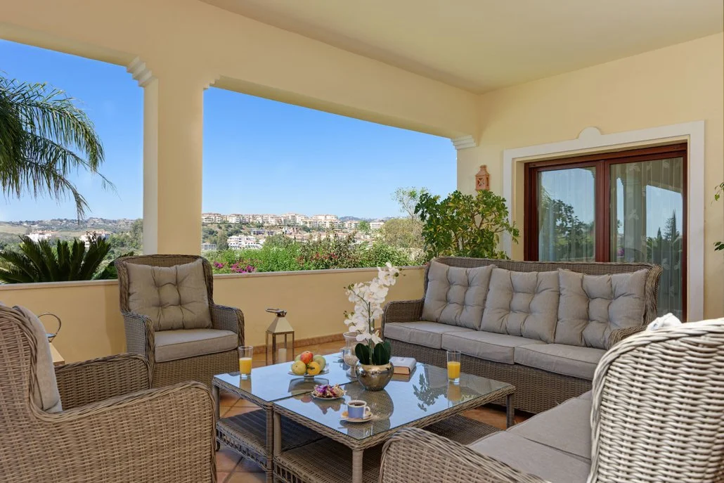 Covered outdoor balcony with wicker furniture, a glass-top coffee table, and a scenic view of a hillside neighborhood and greenery.