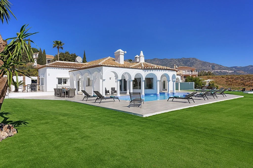 A luxurious white house with a pool and outdoor lounge area, set on a well-maintained grassy yard with mountains in the background.
