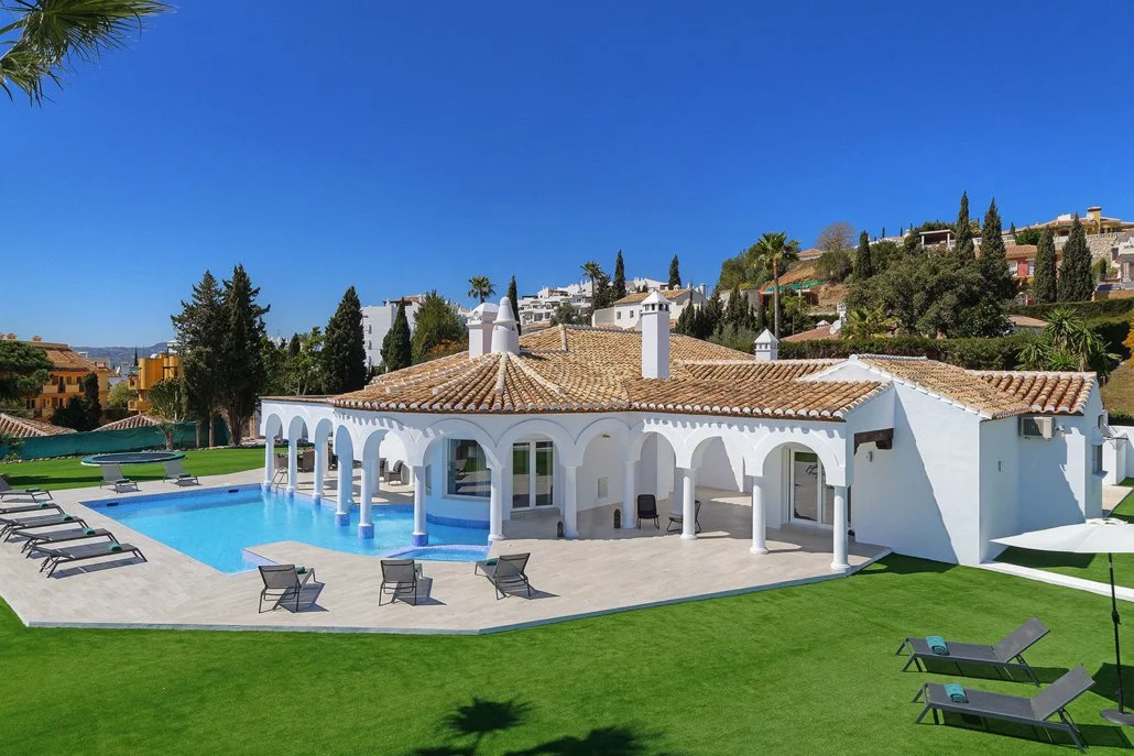 Luxury house with white walls, orange tiled roof, and a large swimming pool with lounge chairs on a sunny day.