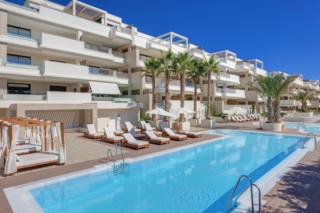 Luxury hotel swimming pool with lounge chairs and palm trees against a modern white building under a clear blue sky.
