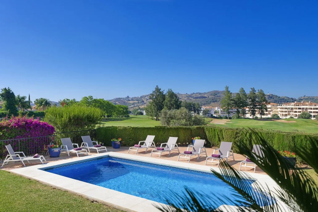 Swimming pool with lounge chairs and colorful flower pots in a sunny outdoor setting of a golf course and distant hills.