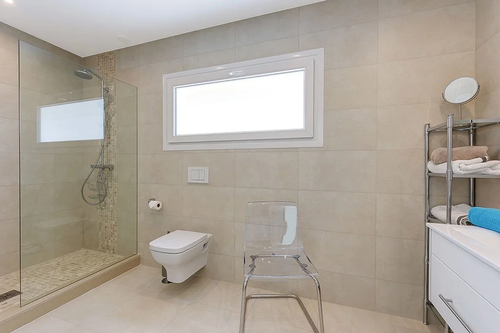 Modern bathroom with walk-in shower, floating toilet, transparent chair, metal shelving with towels, and a large window.