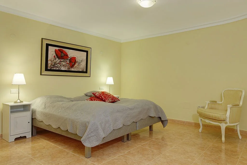 A bedroom with a bed, two bedside lamps, artwork with red flowers above the bed, a white nightstand, and a yellow striped armchair in the corner.