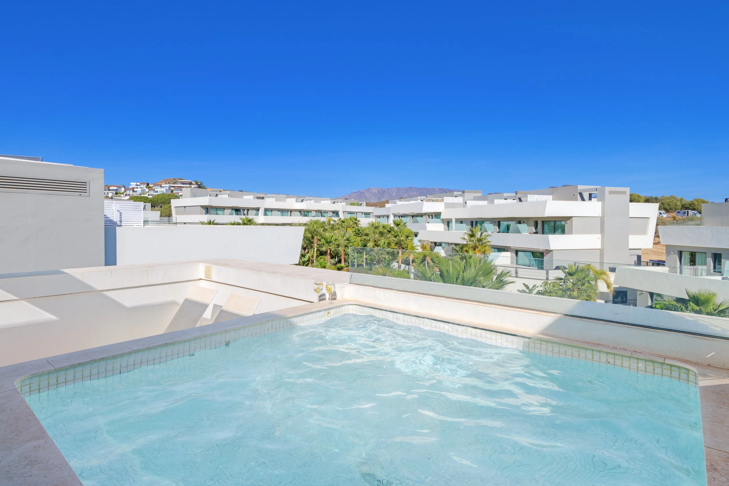 Luxury rooftop swimming pool with modern white residential buildings and palm trees under a clear blue sky.