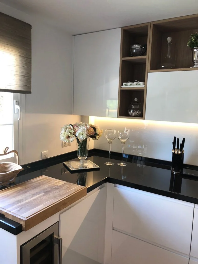 Modern kitchen corner with black countertop, white cabinets, a wooden cutting board, a glass vase with white and pink flowers, two empty wine glasses, and kitchen utensils. There are upper open and closed cabinets and a small window with a brown blin