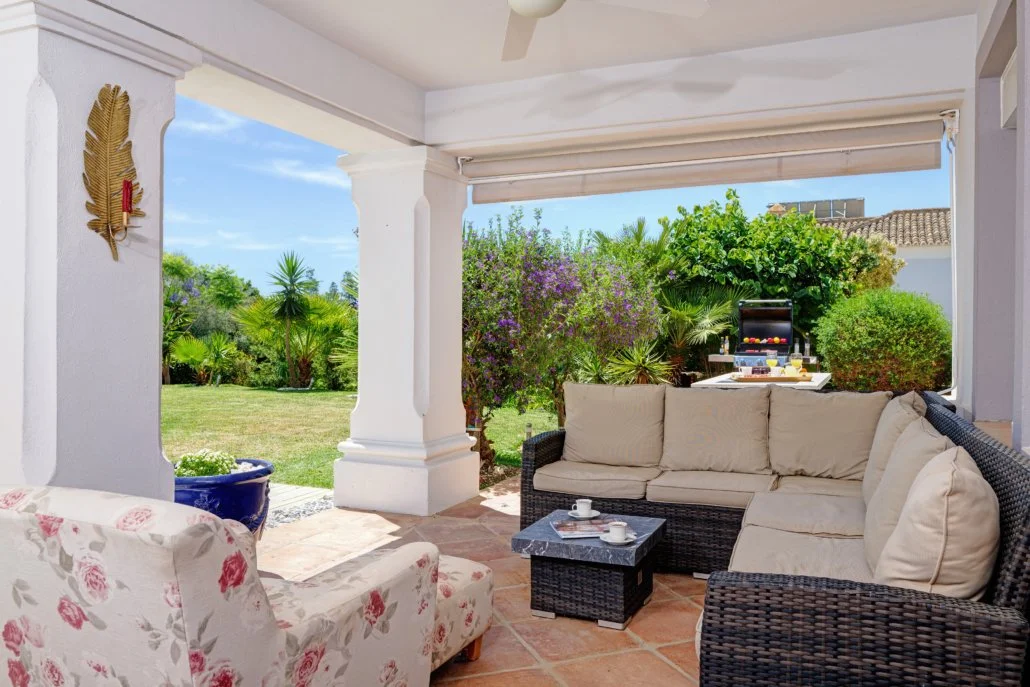 Covered patio with beige cushions, wicker furniture, and a floral armchair overlooking a lush garden with purple and green foliage, and a barbecue grill in the background.