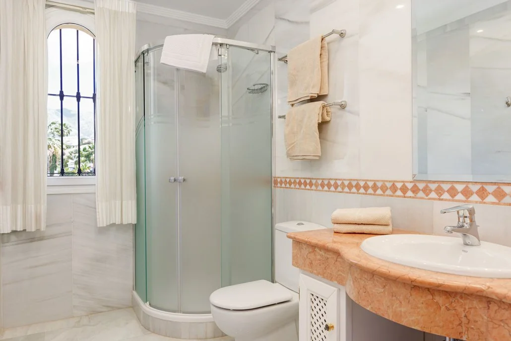 A small, bright bathroom with a frosted glass shower, a window with white curtains, beige towels hanging on a wall, a white toilet, and a beige marble sink with a beige hand towel on top.