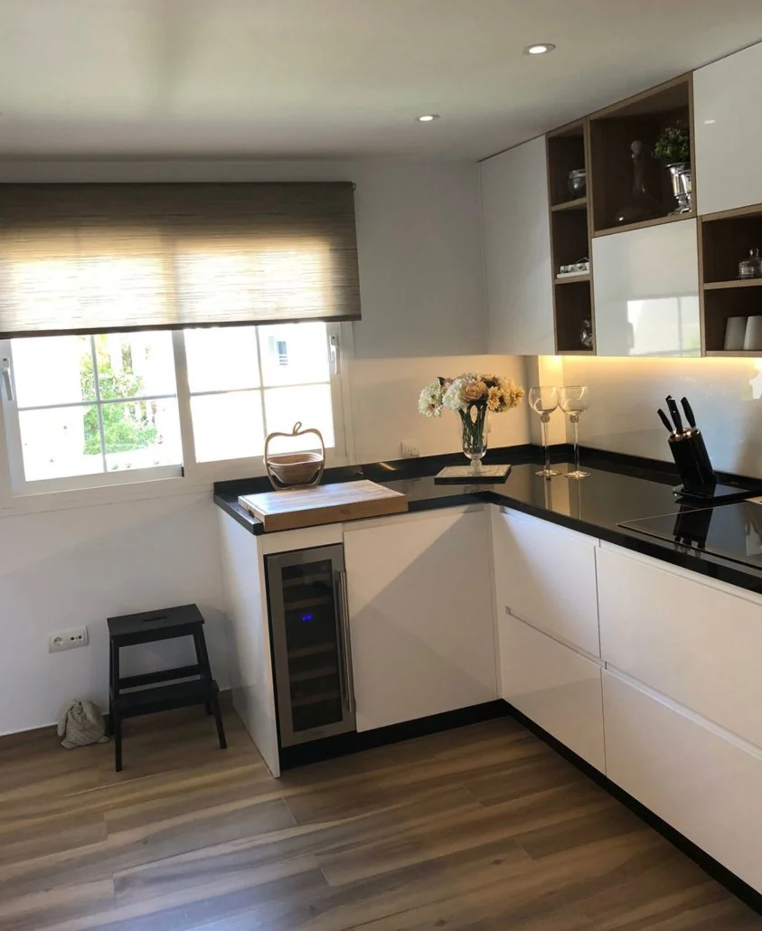 Modern kitchen with white cabinets, black countertops, and open shelving. There is a window with a beige roller blind, a bouquet of flowers, two wine glasses, a knife block, and a small wine fridge. A stool and a cutting board are also present.