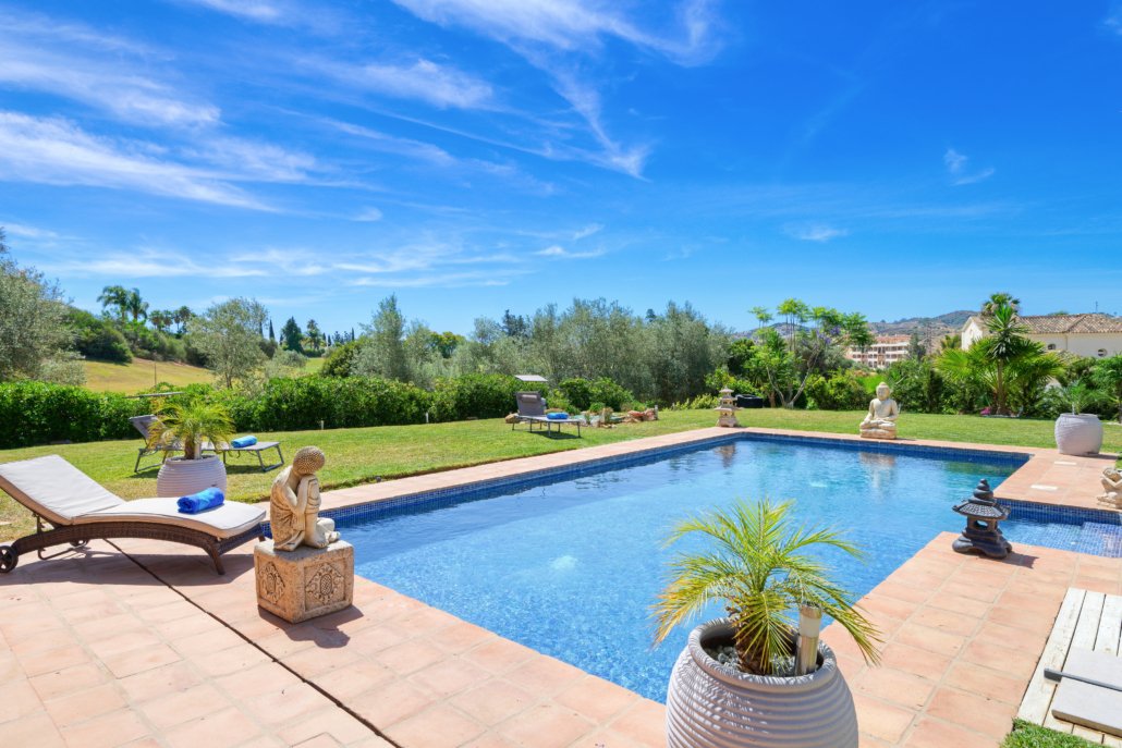 A backyard with a swimming pool, lounge chairs, potted plants, and statues, overlooking a lush green landscape under a clear blue sky.