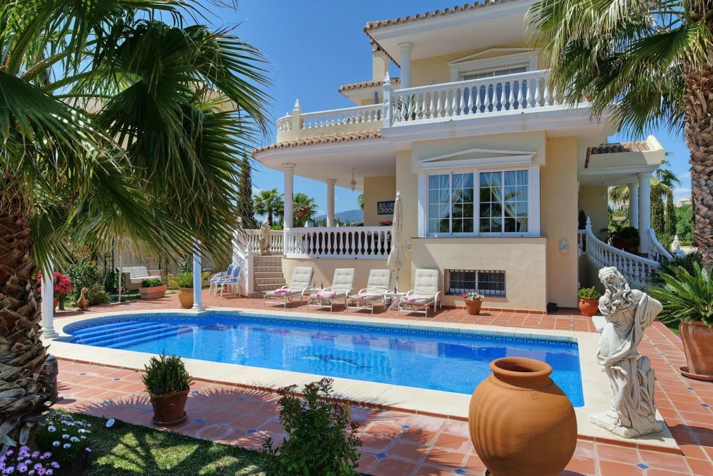 A backyard with a swimming pool, surrounded by potted plants and white lounge chairs, in front of a large, two-story cream-colored house with balconies and decorative columns, set against a blue sky with palm trees.