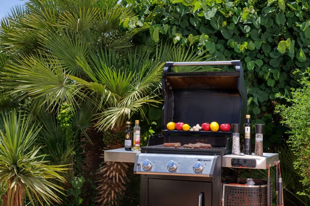 A black gas grill with food and condiments, surrounded by green tropical plants.