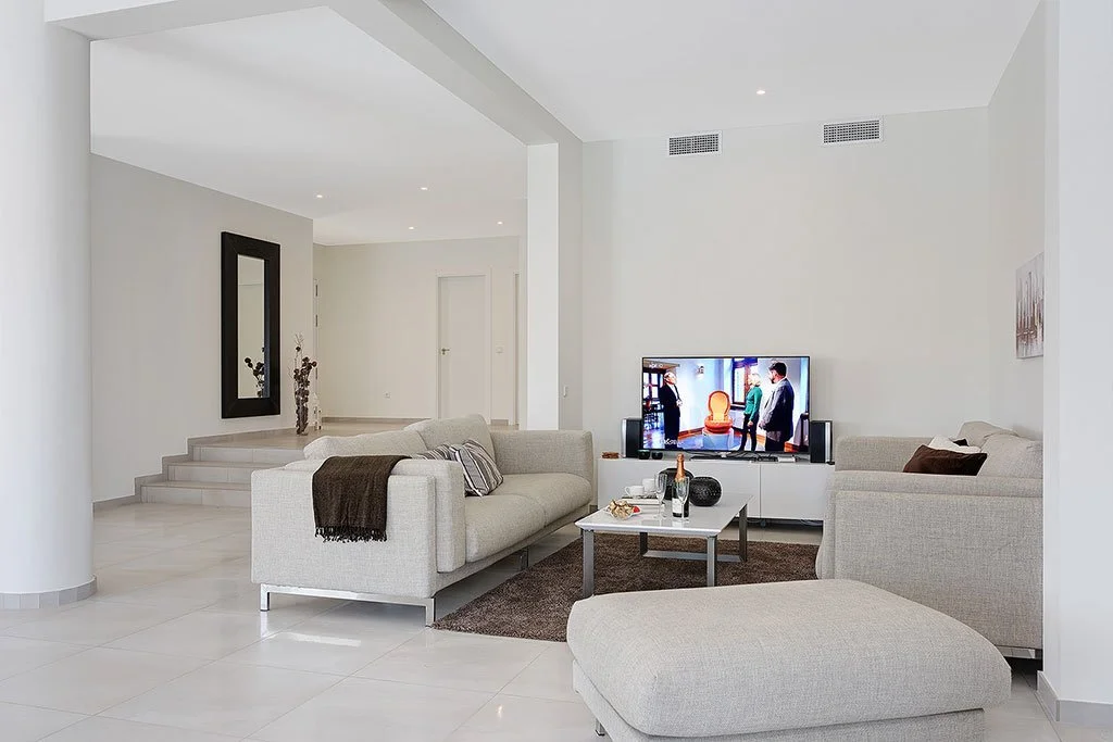 A modern living room with white walls, beige sofas, a flat-screen TV, a coffee table with bottles, glasses, and a decorative item, and a dark brown rug.