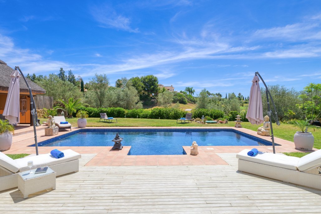 Beautiful backyard with a swimming pool, lounge chairs, umbrellas, lush greenery, and a clear blue sky.