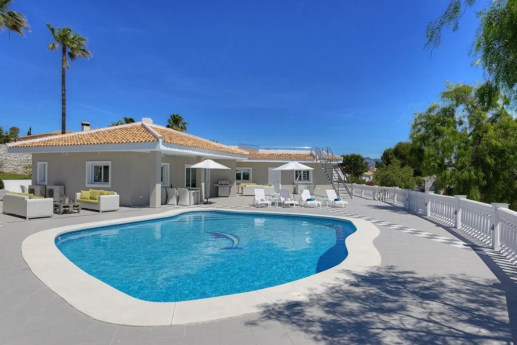 A backyard with a swimming pool, white patio furniture, umbrellas, a house with a tiled roof, and trees under a clear blue sky.