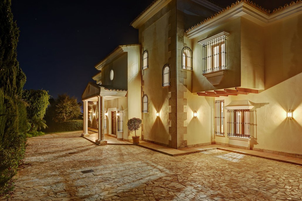 Night view of a large, illuminated house with barred windows and a stone paved driveway.