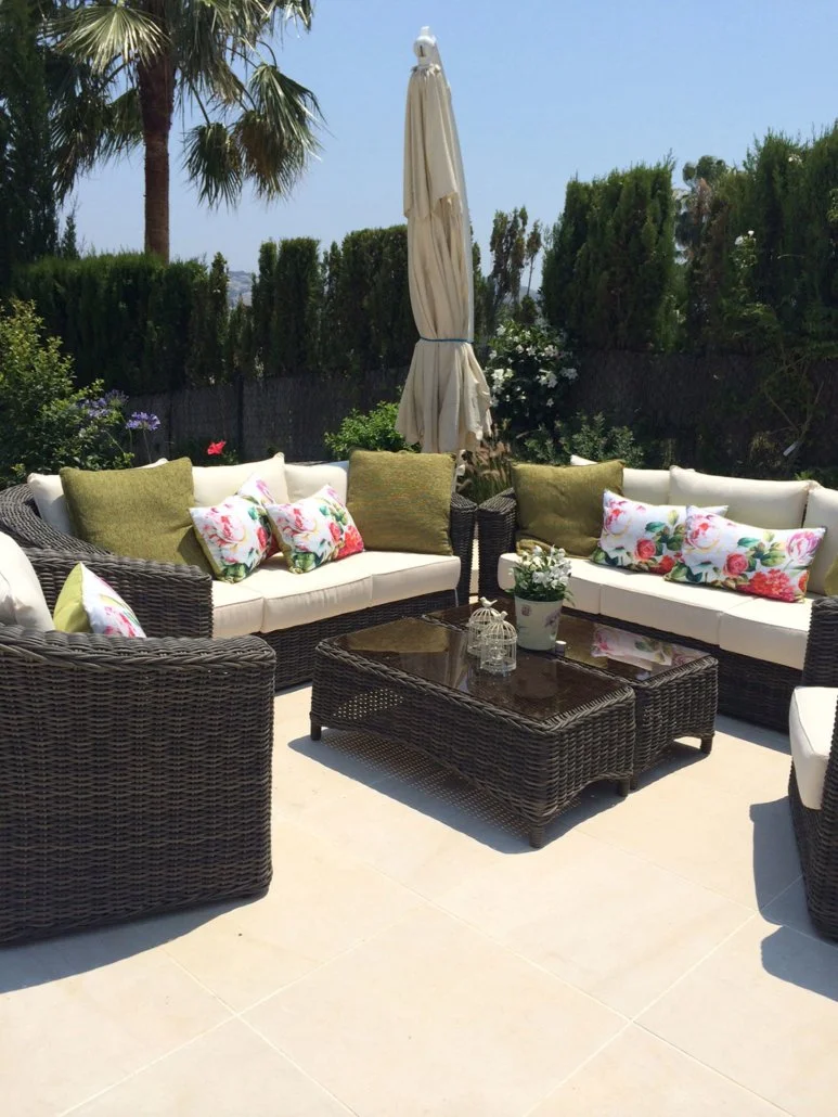 Outdoor patio with wicker sofas and armchairs, floral and green cushions, a glass-top coffee table, potted plants, and a closed beige patio umbrella, surrounded by lush greenery and palm trees under a clear blue sky.