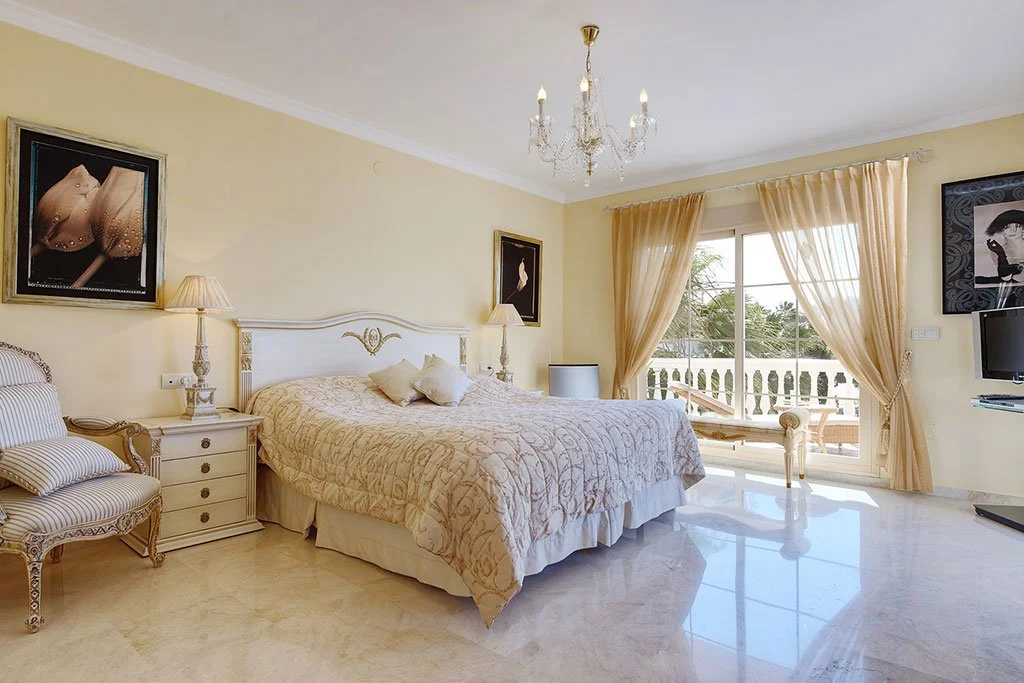 Elegant bedroom with a large bed, cream-colored bedding, and matching curtains, a chandelier hanging from the ceiling, and a balcony with outdoor furniture visible through sliding glass doors.