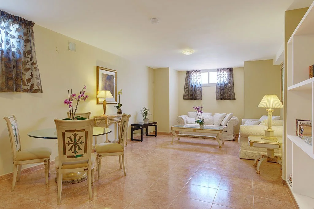 Bright living room with cream-colored sofas, a glass dining table with chairs, and floral curtains.