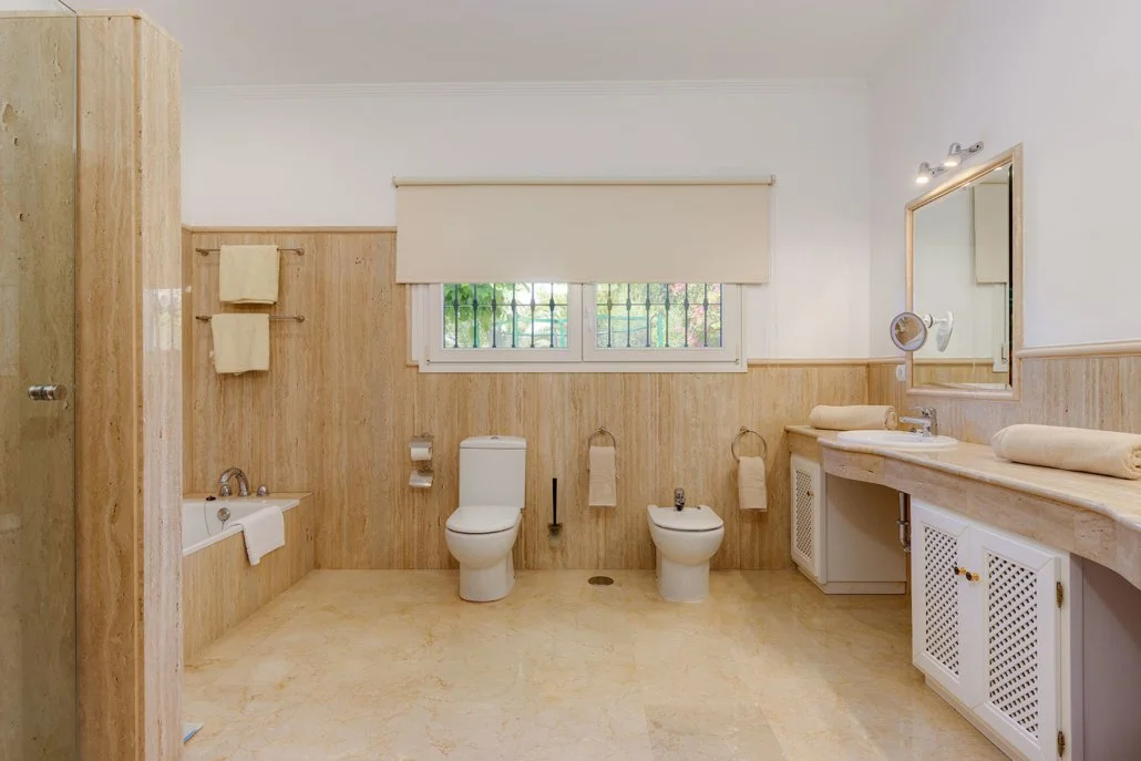 Bathroom with a toilet, bidet, bathtub, large vanity with a mirror, and window with blinds.