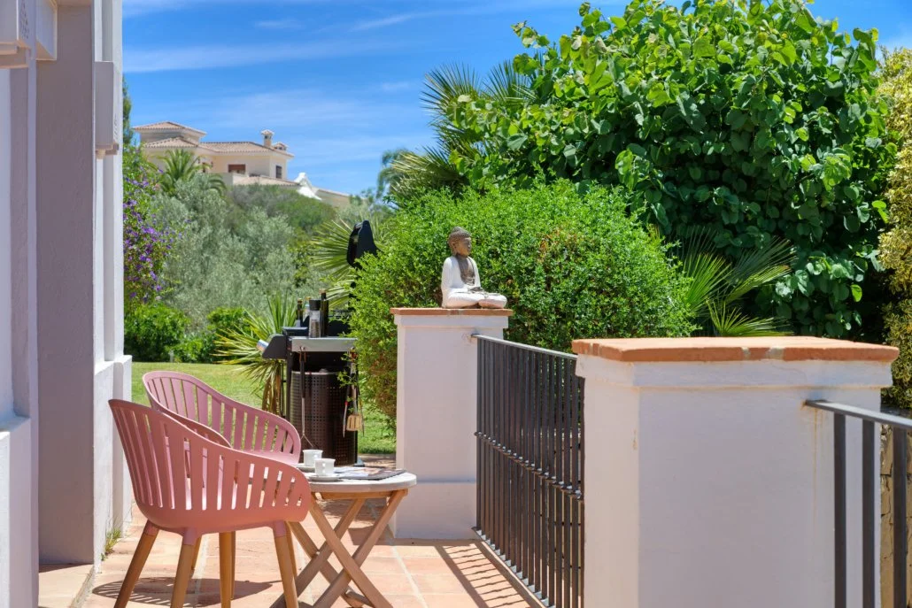 A balcony with two pink outdoor chairs and a small table with cups, overlooking a garden with lush green bushes and trees, and a white building with terracotta roof in the background under a blue sky.