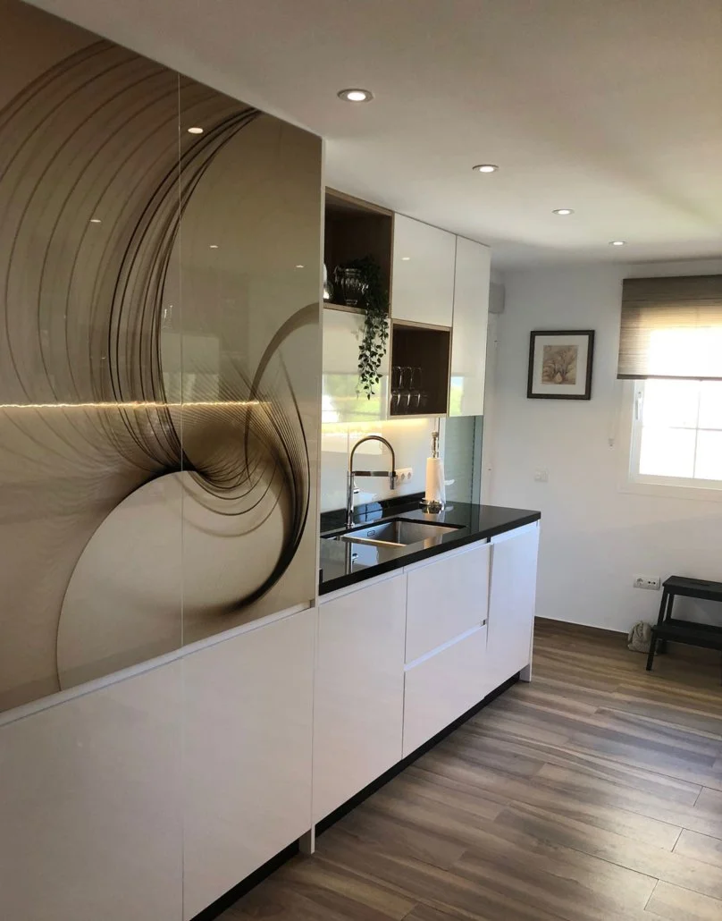 Modern kitchen with white cabinets, black countertop, and a sink with a gold faucet. There is an abstract wall art with swirls and lines on the left side, a window with blinds on the right, and a framed picture on the wall. The floor is wood with a s