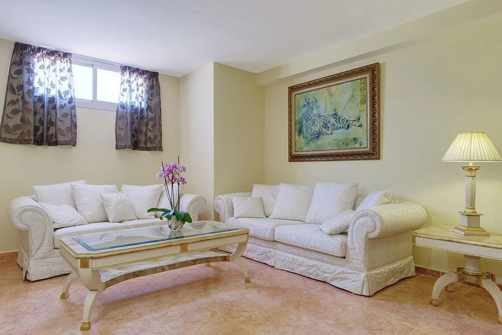 Living room with two white sofas, a glass coffee table with floral decoration, a side table with a lamp, a framed painting on the wall, and a window with floral curtains.