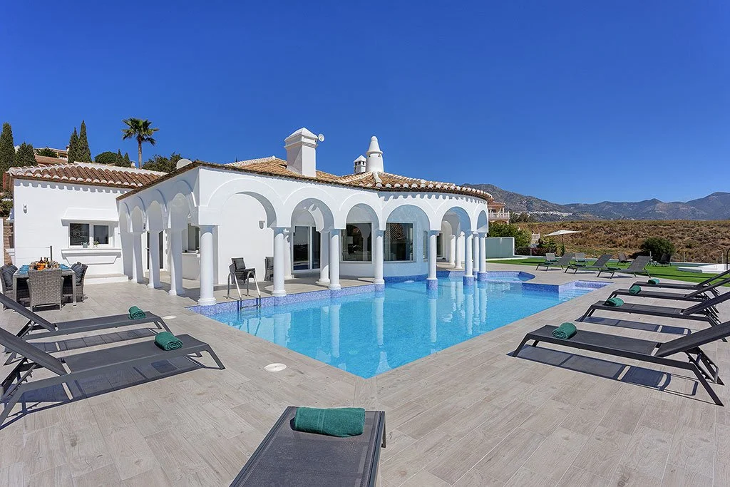 Luxury house with a swimming pool, lounge chairs with green towels, and mountain views.