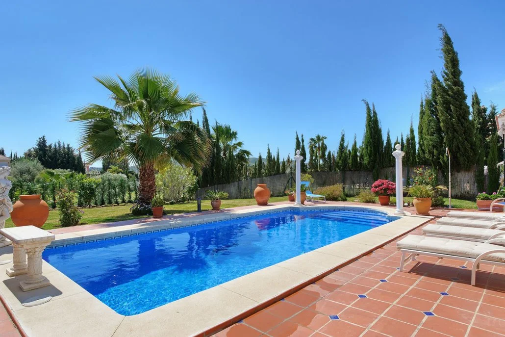 Sunny backyard with a swimming pool surrounded by potted plants, palm trees, and lounge chairs, on a clear day.