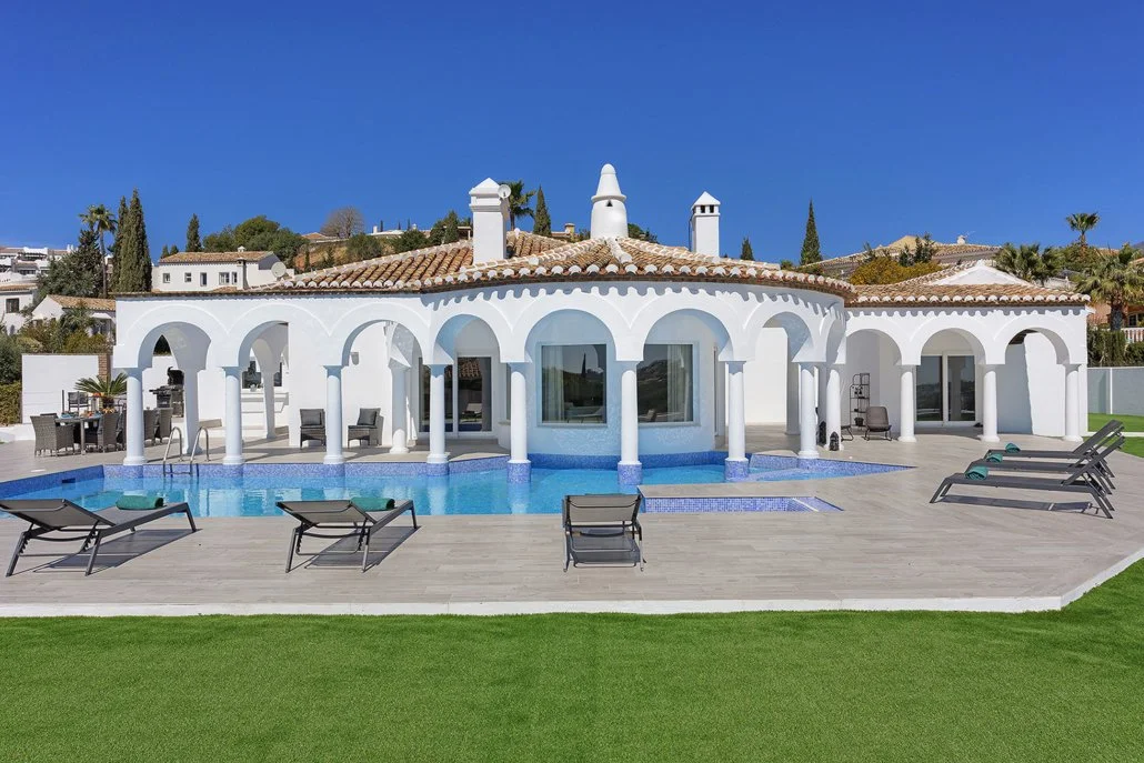 White Mediterranean-style house with arched windows and columns, swimming pool, poolside lounge chairs, and a tiled roof under a clear blue sky.