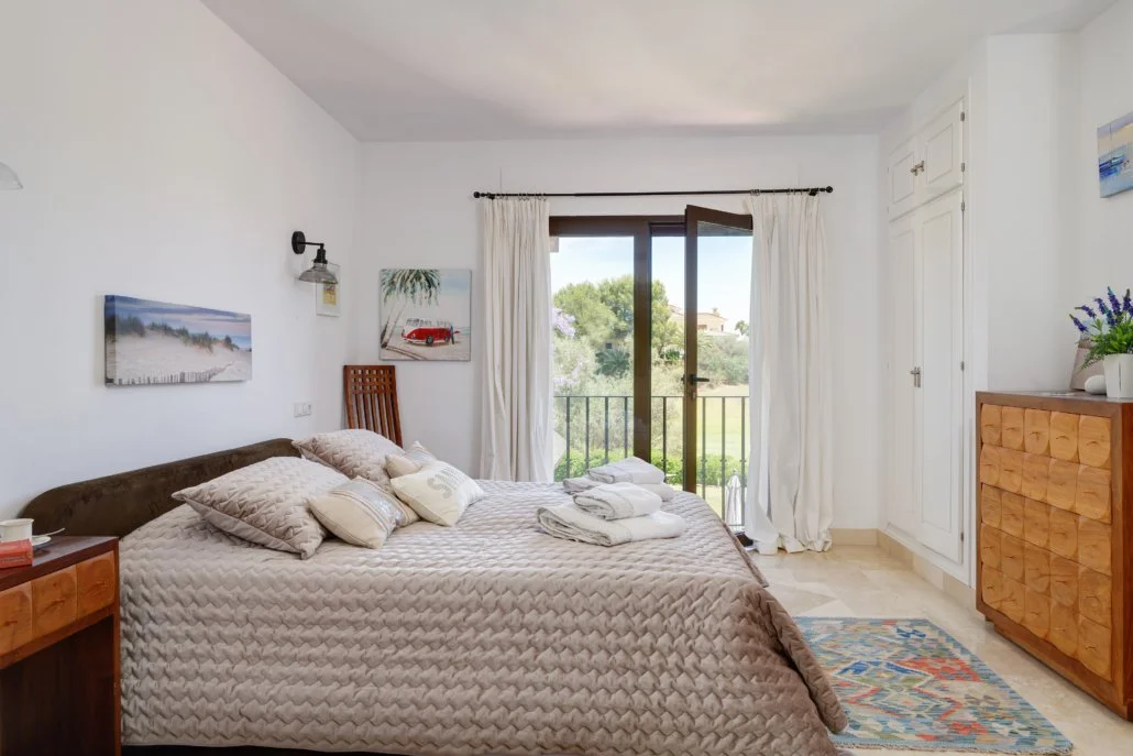 A bedroom with a large bed covered in beige and white pillows, next to a window with white curtains and a sliding glass door leading to a balcony. There are paintings on the wall and a wooden dresser with a plant on top.