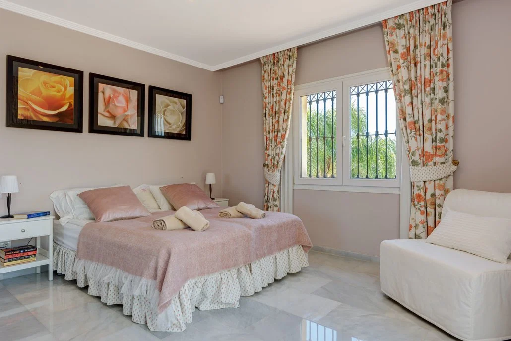 A bedroom with a bed covered in a pink blanket and pillows, a white armchair, two nightstands with lamps, framed floral pictures on the wall, and a window with floral curtains showing greenery outside.