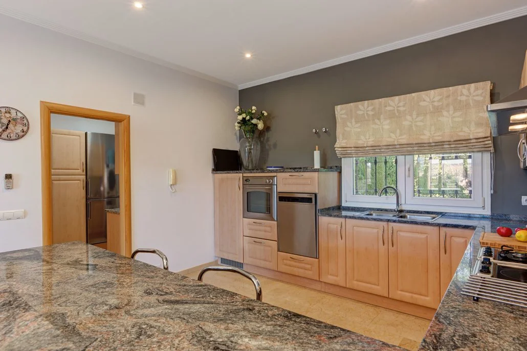 Kitchen with light wooden cabinets, a poster window with a beige roller shade, dark speckled granite countertops, a double sink, a built-in oven, dishwasher, and a stove with colorful peppers. There is a vase with flowers on top of the cabinet and a 