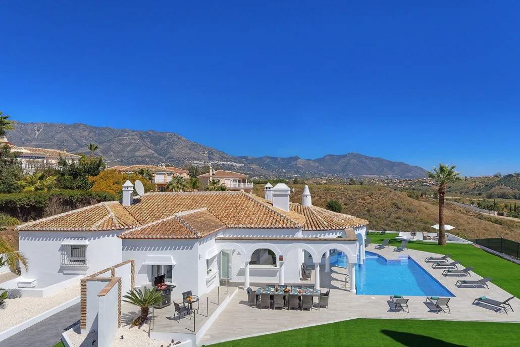 Luxury villa with white walls, terracotta roof, outdoor swimming pool, and lounge chairs, set against mountainous landscape under a clear blue sky.