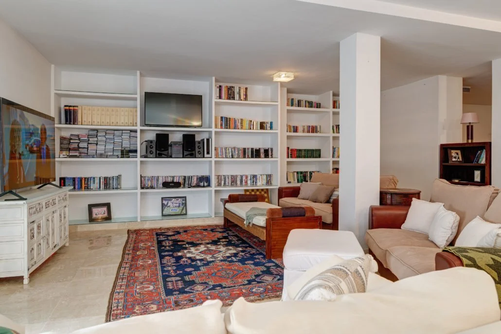 Living room with built-in white bookshelves, a TV, and various books and DVDs. Contains sofas, a patterned area rug, and other furniture.